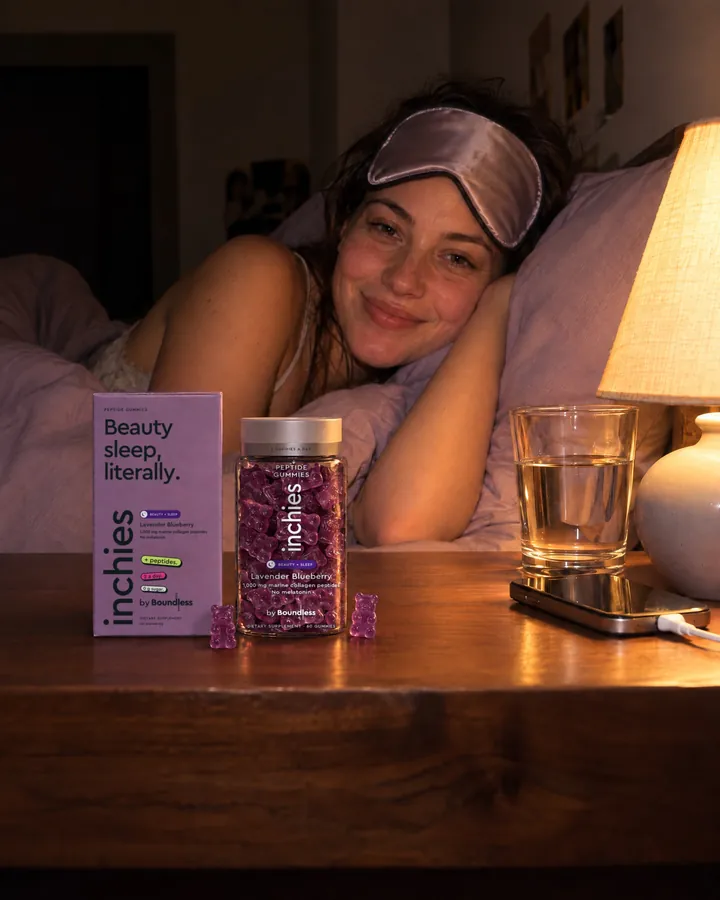 A woman in bed at night with a sleep mask, smiling, a lavender inchies Beauty + Sleep jar on the bedside table