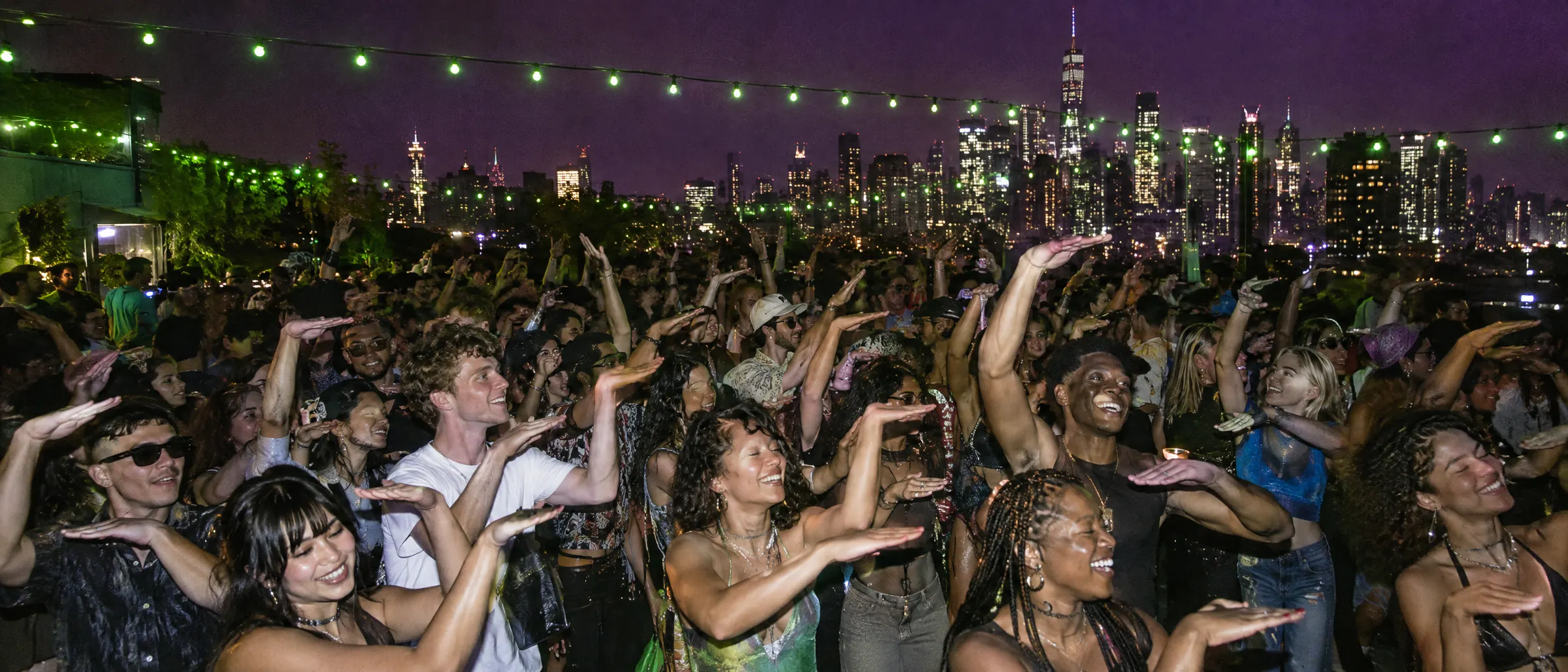 A rooftop block party at night, a crowd of young people making the hand sign under lime string lights